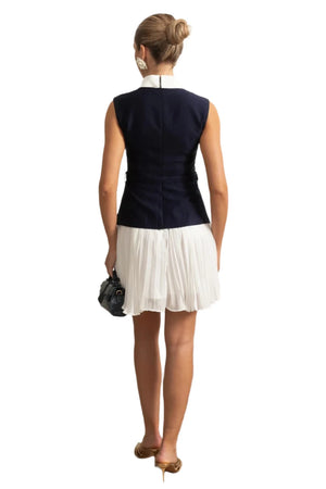 Woman in a navy top and white skirt standing in a room with decorative wall and plant.