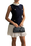Woman in a navy sleeveless top and white skirt holding a black handbag against a decorative wall.