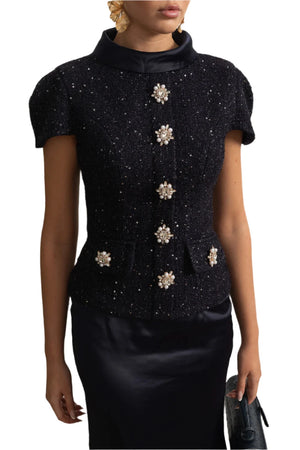 Black top with floral embellishments worn by a person against a decorative wall.