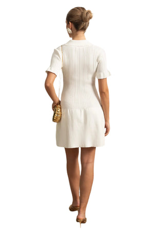Woman in a white dress standing in a stylish room with decorative wall molding and a plant.